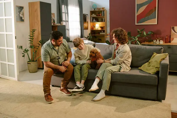 Kafkasyalı çocuk kanepeye oturmuş, küçük kahverengi köpeği okşuyor. Yanında beyaz bir kadın ve orta yaşlı bir adam gülümsüyor. Oturma odasında ilk evcil hayvanı yakalıyor.