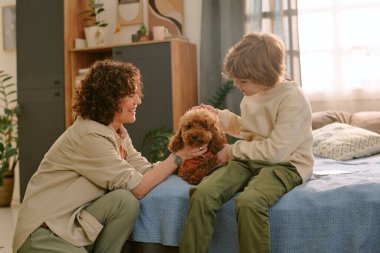 Kafkasyalı orta yaşlı bir kadın yatağın yanında diz çöküp gülümserken beyaz bir çocuk hafifçe küçük kahverengi bir köpeği okşuyor.