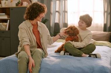 Kafkasyalı orta yaşlı kadın ve beyaz çocuk yatakta oturmuş gülümsüyor ve küçük kahverengi köpeği okşuyor, çocuk evdeki ilk evcil hayvana şefkat gösteriyor, doğal ortam.