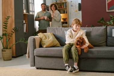 Kafkas çocuk koltukta oturmuş gülümserken küçük kahverengi köpeği okşuyor, beyaz orta yaşlı adam ve kadın arka planda yan yana durup evdeki ilk evcil hayvanla çocuk etkileşimini izliyorlar.