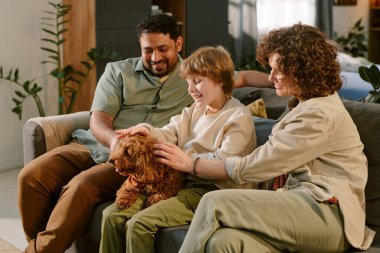 Kafkas kadın, beyaz çocuk ve Orta Doğulu adam koltukta oturmuş küçük kahverengi köpeği okşuyor, çocuk gülümsüyor ve evdeki ilk evcil hayvanla iletişim kuruyor, aile bağı kurma anı.