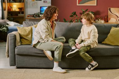 Kafkasyalı genç bir kadın kanepeye oturmuş, gülümsüyor ve beyaz bir çocukla konuşuyor. Bacakları çapraz oturuyor. İkisi de oturma odasında ilk evcil hayvan deneyimi sırasında iletişim kuruyor.