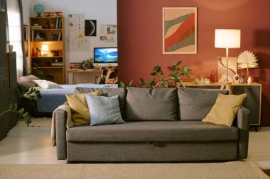 Ön planda gri kanepesi olan boş oturma odası, rahat yatak odasını ve bilgisayar monitörlü çalışma alanını gösteren arka plan, modern apartmanı dekore eden kapalı tesisler.