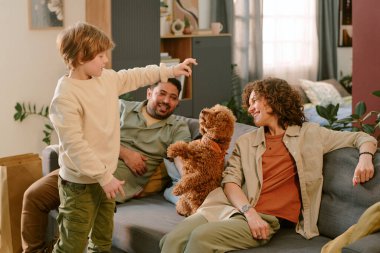 Kafkas çocuk küçük kahverengi köpekle oynarken orta yaşlı beyaz kadın ve orta yaşlı adam kanepede izliyor ve gülümsüyor, ev tecrübelerinde ilk evcil hayvanı yakalıyor.