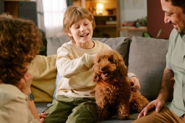 Kafkas çocuk gülümseyerek iki yetişkin arasındaki koltukta oturan tüylü kahverengi köpeği okşuyor ve evdeki ilk evcil hayvan deneyimi sırasında neşeli bir etkileşim gösteriyor.
