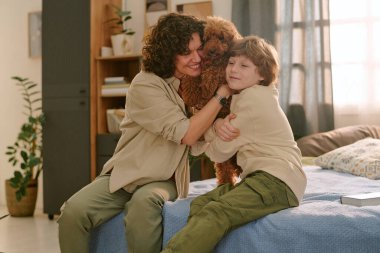 Kafkasyalı orta yaşlı kadın ve beyaz çocuk yatakta oturmuş küçük kahverengi köpeğe sarılıyor ve gülümsüyor.