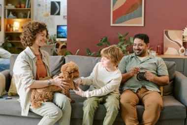 Beyaz bir kadın küçük bir köpeği tutarken, beyaz bir çocuk köpeğini okşuyor, Orta Doğulu bir adam gülümsüyor, elinde kupa tutuyor, evde ilk evcil hayvanla vakit geçiriyor.