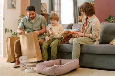 Orta yaşlı beyaz bir adam alışveriş çantalarını boşaltırken beyaz bir çocuk koltukta oturuyor ve Orta yaşlı beyaz bir kadın izliyor, küçük bir köpek çocuğun yanında yatıyor, evcil hayvan malzemeleri görünür.