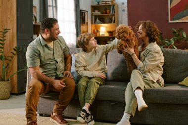 Beyaz bir çocuk orta yaşlı beyaz bir kadın ve orta yaşlı bir adamla koltukta oturuyor. Gülümsüyor ve pofuduk bir köpeği okşuyor. Aile evdeki ilk evcil hayvanla etkileşimde.