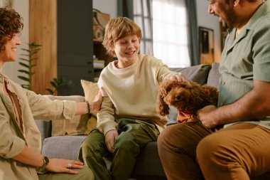 Beyaz çocuk kanepeye oturmuş gülümserken küçük bir köpeği okşuyor. Etrafı orta yaşlı beyaz kadın ve orta yaşlı adamla çevrili. Ailesi evdeki ilk evcil hayvanla etkileşimde.