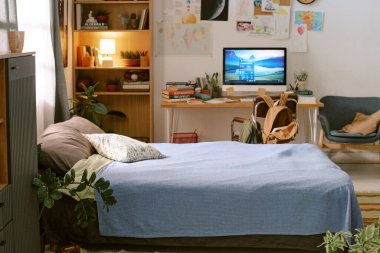 Rahat yatak odası sahnesi, yeşil ev bitkileriyle çevrili mavi battaniyeli temiz bir yatak odası, bilgisayar monitörü ve sırt çantalı çalışma masası, ev ortamındaki ilk evcil hayvanı gösteriyor.