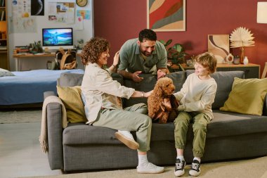 Kafkasyalı bir çocuk kanepeye oturup küçük kahverengi bir köpeği okşar. Bu sırada farklı ebeveynleri gülümser ve evcil hayvanlarla etkileşime geçer.