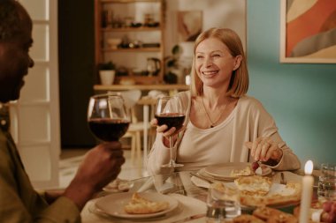 Üst düzey beyaz kadın, son sınıf siyahi bir adamla yemek masasında otururken gülümsüyor, elinde şarap bardakları, birlikte yemek yiyor, sıcak bir yuva kuruyor, romantizm ve arkadaşlığı ifade ediyor.