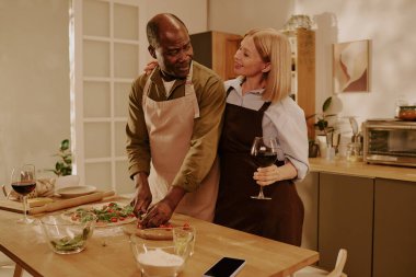 Kıdemli siyahi adam salata hazırlarken yanında şarap kadehi tutan kıdemli beyaz kadın hem gülümsüyor hem de mutfak ortamında sevgi dolu bir anı paylaşıyor.