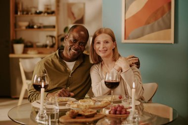 Kıdemli siyah adam ve kıdemli beyaz kadın birlikte yemek masasında oturuyor gülümsüyor ve sarılıyor, şarap ve yemekle romantik bir akşam yemeğinin tadını çıkarıyor, mutlu ve bağlı görünüyor.