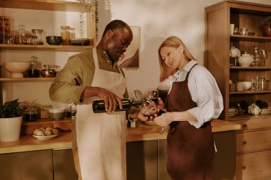 Üst düzey siyahi adam, mutfakta kıdemli beyaz bir kadının elinde bulunan bardağa şarap dolduruyor. Önlük takıyor ve gülümsüyor.