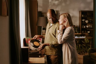 Üst düzey siyahi adam plak plağı tutarken, kıdemli beyaz kadın onu yakından kucaklıyor, ikisi de kapalı mekanda romantik bir an paylaşıyor.