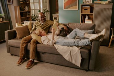 Üst düzey beyaz kadın, kafası yaşlı siyahi adamın kucağında kanepede uzanıyor, gülümsüyor, dijital tableti tutarken birlikte dinleniyor, evde romantik bir an yaşıyor.