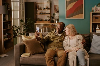 Kıdemli siyahi adam ve kıdemli beyaz kadın kanepede oturup selfie çekiyorlar, gülümsüyorlar ve kucaklaşıyorlar, samimi oturma odasında romantik bir bağ kuruyorlar