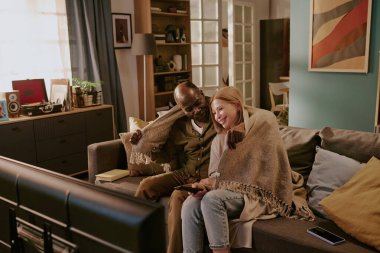 Kıdemli siyah adam ve kıdemli beyaz kadın kanepede yan yana oturuyor, televizyon izlerken gülümsüyor ve battaniyeyi paylaşıyor, rahat ev ortamında romantik bir bağ kuruyor.