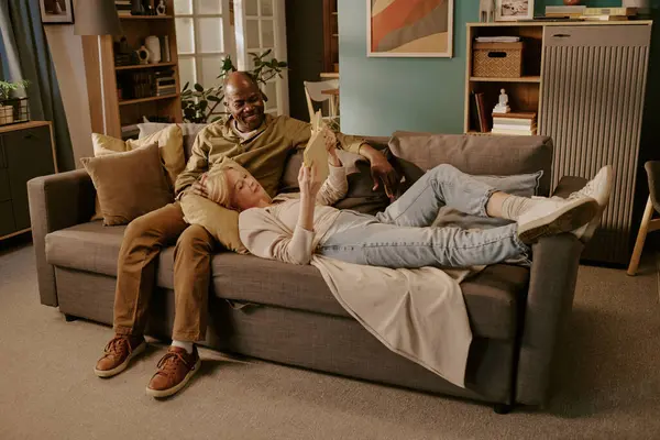 Üst düzey beyaz kadın, kafası yaşlı siyahi adamın kucağında kanepede uzanıyor, gülümsüyor, dijital tableti tutarken birlikte dinleniyor, evde romantik bir an yaşıyor.