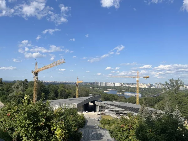 市内の川岸のグリーンゾーンに工業クレーンを設置した建設現場の様子 昼間の雲で青空 ストック画像