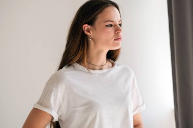 Beyaz pamuklu bir tişörtün içinde ciddi bakışları olan güzel bir genç kadın. Çeşitli temel giysiler giyen bir kadın. Model yap. Fikirleri kabul et. Giysileri kapatın. Asgari düzeyde. Basitlik.