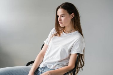 Beyaz pamuklu tişört ve açık mavi kot giyen, uzun saçlı, makyözlü, sıradan bir sandalyede oturan güzel bir kadın. Model yap. Fikirleri kabul et. Giysileri kapatın. Asgari düzeyde. Basitlik.