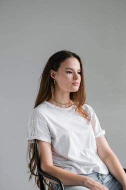 Beyaz pamuklu tişörtlü, uzun saçlı, makyözlü, sıradan bir sandalyede oturan güzel bir kadın. Çeşitli temel giysiler giyen bir kadın. Model yap. Fikirleri kabul et. Giysileri kapatın. Asgari düzeyde. Basitlik.