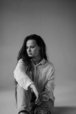 Yüzünde üzgün ve ciddi bir ifadeyle yerde oturan uzun beyaz gömlekli ve kot pantolonlu güzel bir kadın. Duygularını ve duygularını ifade eden güzel bir kadın..