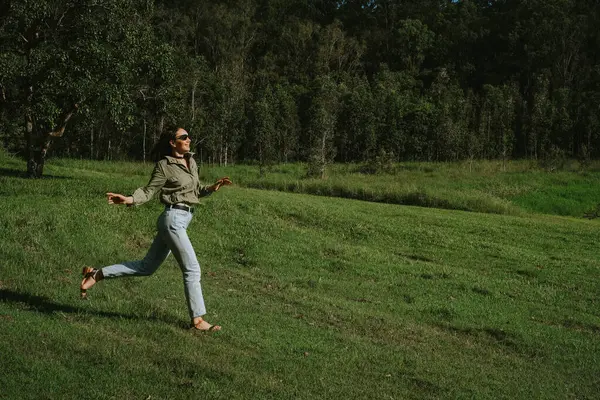 Yeşil elbiseli, mavi kotlu, uzun saçlı bir kadın çayırda koşuyor ve doğanın tadını çıkarıyor.