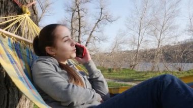 Genç ve güzel bir kız doğada bir hamakta dinleniyor ve akıllı bir telefonla konuşuyor..