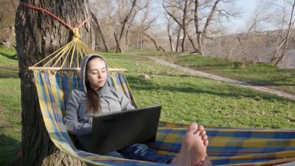 ノートパソコンを持つハンモックの少女は 晴れた日には自然にリモートで動作します リモートコミュニケーションと仕事のフリーランスの概念 — ストック動画