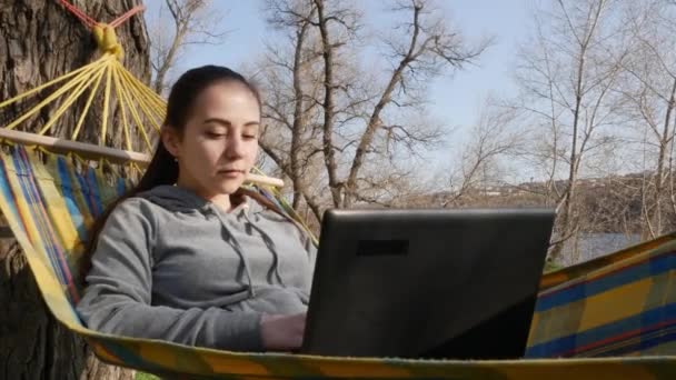 ノートパソコンを持つハンモックの少女は 晴れた日には自然にリモートで動作します リモートコミュニケーションと仕事のフリーランスの概念 — ストック動画