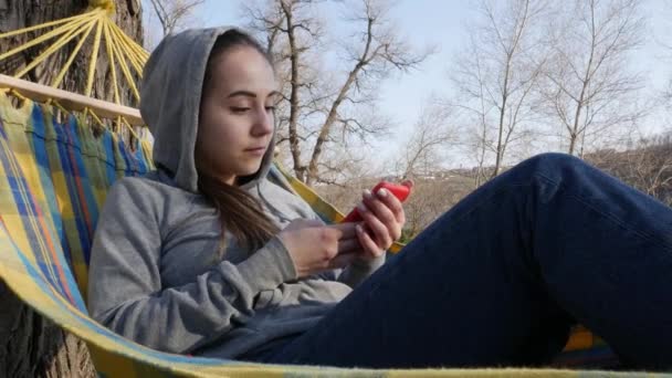 美しい若い女の子は自然の中でハンモックで休んでいて スマートフォンで話しています — ストック動画