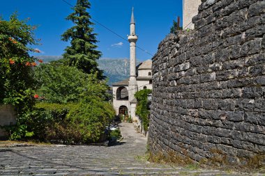 Ön planda sağlam bir tuğla duvar dimdik dururken, arka planda Cirokastra 'daki camiye ait uzun ve zarif bir minare gökyüzüne ulaşıyor.