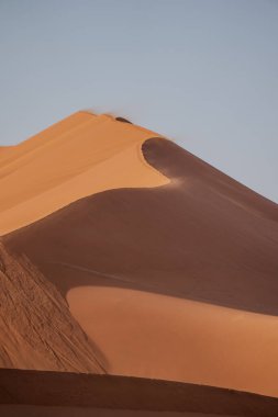 Rüzgar, Sossusvlei, Namibya 'da gün doğumunda dev bir kum tepesinin üzerinden kum üflüyor.