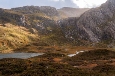 Güneş ışığı, sonbaharda Galler, Snowdonia 'da bir gölün üstündeki bir dağın tepesinde bulutların arasından doğuyor.