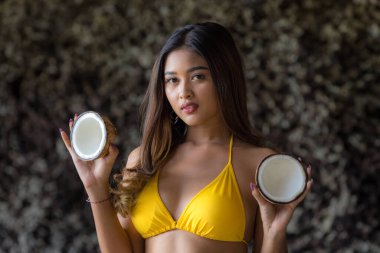 Hindistancevizi tutan ve mayo giyen çekici Asyalı kadın portresi. Kayanın yanında duran güzel kadın. Bronzlaşmış deri. Yaz kavramı. Tropikal adaya git. Suluban Sahili, Bali, Endonezya