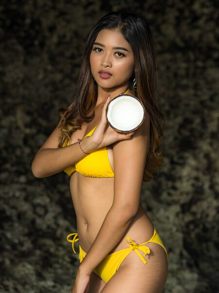 Привлекательная азиатка с кокосом и в жёлтом купальнике. Улыбающаяся женщина, стоящая у скалы. Загорелая кожа. Летняя концепция. Поездка на тропический остров. Пляж Сулубан, Бали, Индонезия