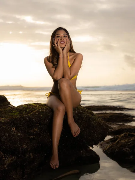 年轻的亚洲女人穿着比基尼坐在岩石上 性感的身体 把日落时间花在海滩上 热带岛屿的暑假 旅行生活方式 垂直布局 Suluban海滩 印度尼西亚巴厘 — 图库照片
