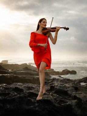 Kumsalda kemanı olan güzel bir kadın. Müzik ve sanat konsepti. Kısa kırmızı elbise giyen ve doğada keman çalan zayıf bir kız. Gün batımı zamanı. Bulutlu gökyüzü. Bali, Endonezya