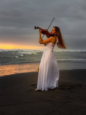 Kumsalda kemanı olan beyaz bir kadın. Müzik ve sanat konsepti. İnce kız uzun beyaz elbise giyiyor ve doğada keman çalıyor. Gün batımı zamanı. Bulutlu gökyüzü. Bali, Endonezya