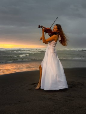 Kumsalda kemanı olan beyaz bir kadın. Müzik ve sanat konsepti. İnce kız uzun beyaz elbise giyiyor ve doğada keman çalıyor. Gün batımı zamanı. Bulutlu gökyüzü. Bali, Endonezya