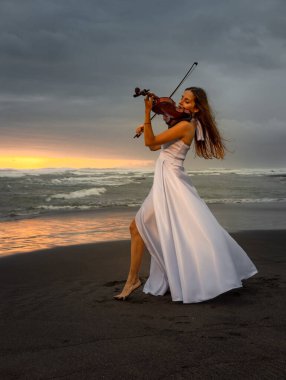 Kumsalda kemanı olan beyaz bir kadın. Müzik ve sanat konsepti. İnce kız uzun beyaz elbise giyiyor ve doğada keman çalıyor. Gün batımı zamanı. Bulutlu gökyüzü. Bali, Endonezya