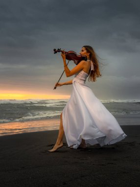 Kumsalda kemanı olan çekici bir kadın. Müzik ve sanat konsepti. İnce kız uzun beyaz elbise giyiyor ve doğada keman çalıyor. Gün batımı zamanı. Bulutlu gökyüzü. Bali, Endonezya