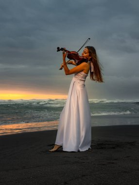 Kumsalda kemanı olan çekici bir kadın. Müzik ve sanat konsepti. İnce kız uzun beyaz elbise giyiyor ve doğada keman çalıyor. Gün batımı zamanı. Bulutlu gökyüzü. Bali, Endonezya