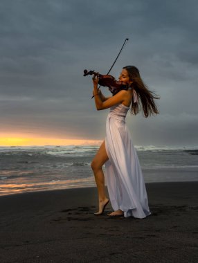 Kumsalda kemanı olan çekici bir kadın. Müzik ve sanat konsepti. İnce kız uzun beyaz elbise giyiyor ve doğada keman çalıyor. Gün batımı zamanı. Bulutlu gökyüzü. Bali, Endonezya