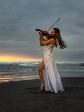 Beautiful woman with violin on the beach. Music and art concept. Slim girl wearing long white dress and playing violin in nature. Sunset time. Cloudy sky. Bali, Indonesia