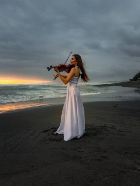 Beautiful woman with violin on the beach. Music and art concept. Slim girl wearing long white dress and playing violin in nature. Sunset time. Cloudy sky. Bali, Indonesia